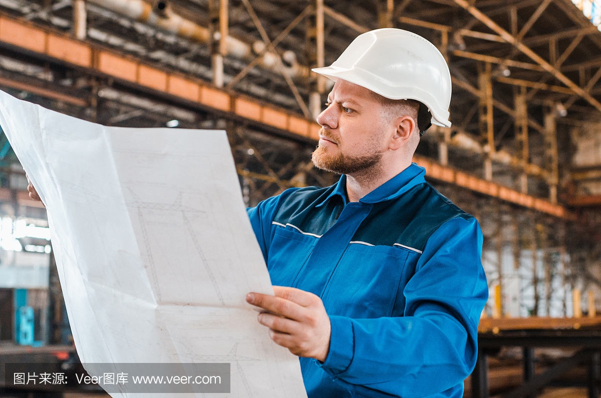 一個穿著工作服、戴著白色頭盔的工人正在檢查一個金屬工廠背景上的圖紙。建筑工人身體很好。男工程師